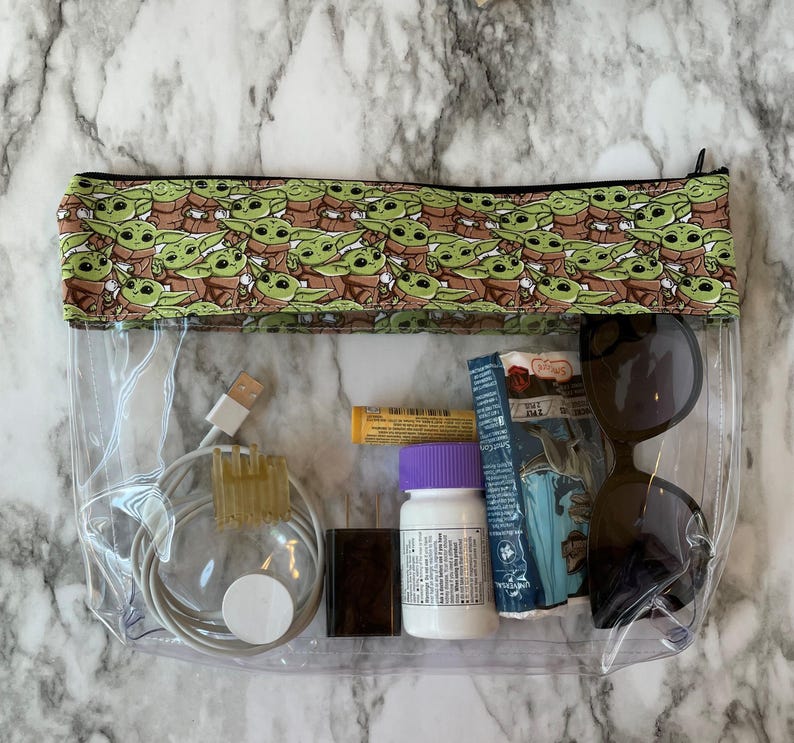 Puede incluir: Una bolsa transparente de pl&aacute;stico con cremallera, con una cremallera negra y un forro de tela verde y marr&oacute;n con un patr&oacute;n repetido de personajes de dibujos animados de Baby Yoda. La bolsa contiene un cable de carga blanco, una peque&ntilde;a botella de pastillas, un peque&ntilde;o tubo amarillo, un objeto rectangular negro y unas gafas de sol negras.