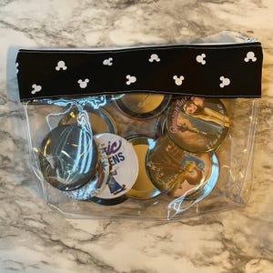Pu&ograve; includere: Una pochette trasparente con cerniera in plastica con una patta in tessuto nero con orecchie di Topolino bianche. La pochette contiene una collezione di spille rotonde con vari disegni colorati, tra cui una spilla con il testo "Magic Queens" e un'altra con il testo "C'era una volta".