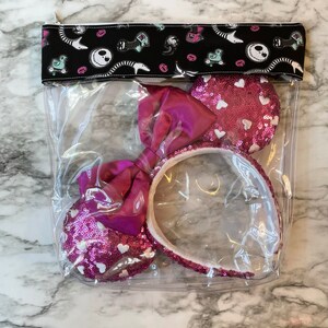 May include: A clear plastic bag with a black and white fabric zipper closure featuring a cartoon character. Inside the bag is a pair of pink Minnie Mouse ears with white sequins and a pink satin bow.