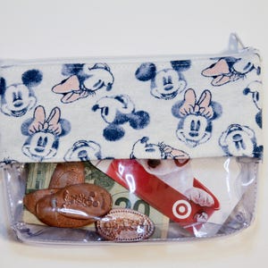 May include: A clear plastic zippered pouch with a white fabric lining featuring a repeating pattern of Mickey Mouse and Minnie Mouse. The pouch is filled with various items, including coins, a gift card, and cash.