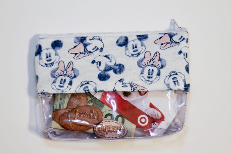 May include: A clear plastic zippered pouch with a white fabric lining featuring a repeating pattern of Mickey Mouse and Minnie Mouse. The pouch is filled with various items, including coins, a gift card, and cash.