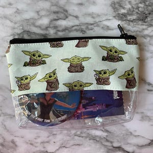 May include: A clear plastic zippered pouch with a green and white fabric lining. The fabric lining has a repeating pattern of a cartoon character with large eyes and green skin. The pouch is filled with various items, including a round sticker with the text "For the first time in forever!" and a card with the text "Disneyland".