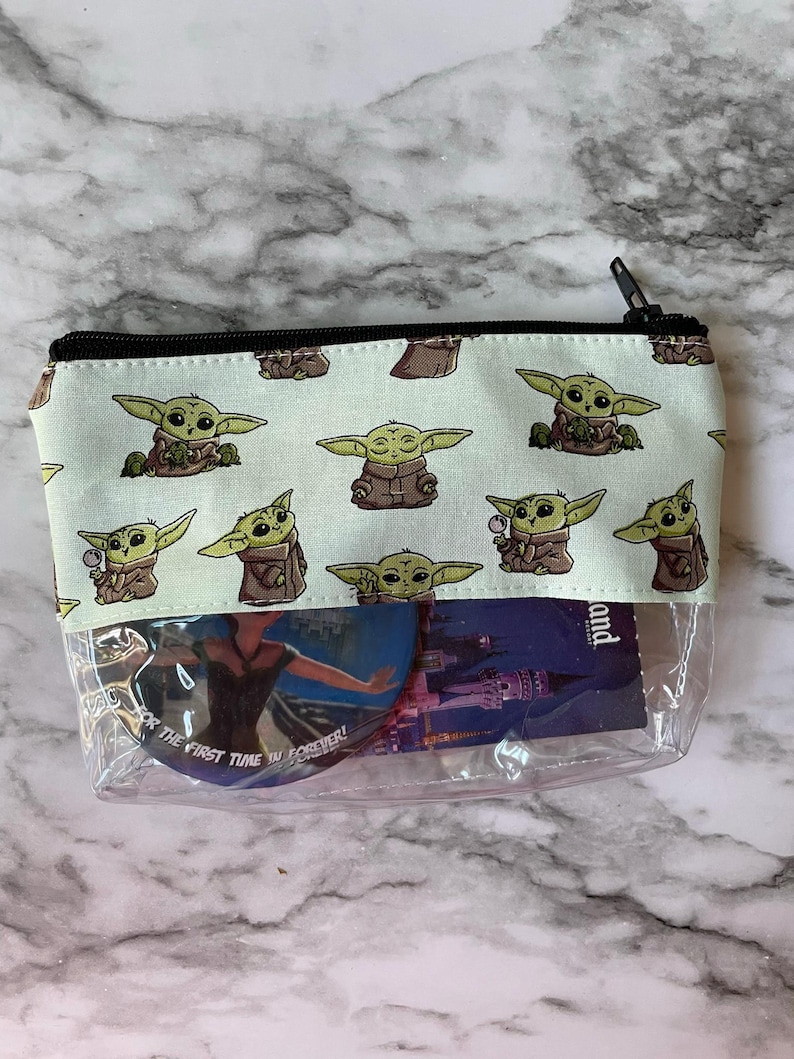 May include: A clear plastic zippered pouch with a green and white fabric lining. The fabric lining has a repeating pattern of a cartoon character with large eyes and green skin. The pouch is filled with various items, including a round sticker with the text "For the first time in forever!" and a card with the text "Disneyland".