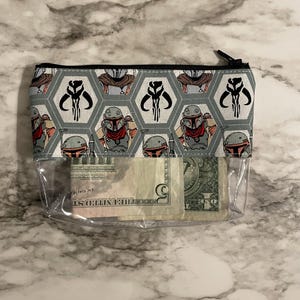 Op de afbeelding: Een doorzichtige, met een rits afsluitbare tas met een grijze en zwarte stoffen voering. De stoffen voering heeft een herhalend patroon van een Mandalorian helm en het symbool van de Mandalorian clan. De tas bevat een aantal Amerikaanse dollarbiljetten.