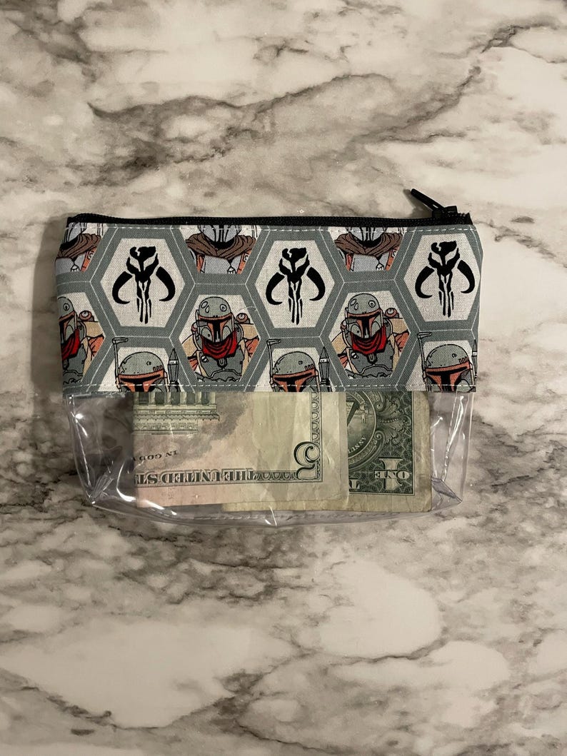 Op de afbeelding: Een doorzichtige, met een rits afsluitbare tas met een grijze en zwarte stoffen voering. De stoffen voering heeft een herhalend patroon van een Mandalorian helm en het symbool van de Mandalorian clan. De tas bevat een aantal Amerikaanse dollarbiljetten.