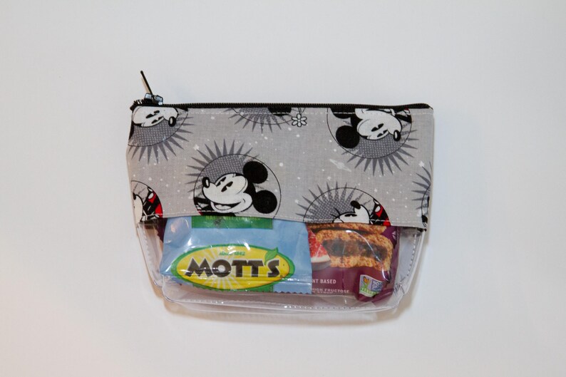 May include: A grey zippered pouch with a Mickey Mouse print. The pouch is clear and contains a Mott's fruit snacks packet.