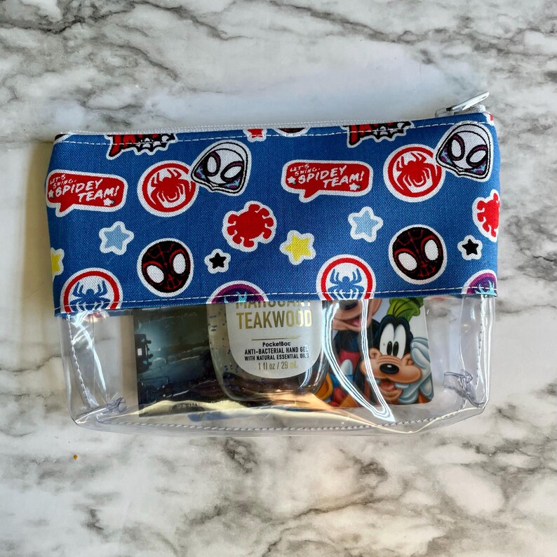 May include: A clear plastic zippered pouch with a blue fabric lining featuring a repeated pattern of Spider-Man characters and the text "Let's Go Spidey Team!" The pouch contains a small bottle of hand sanitizer and a card with a cartoon character.