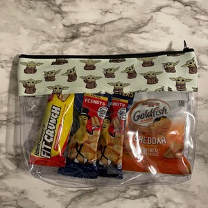 May include: A clear plastic zippered pouch with a green and black fabric lining featuring a repeating pattern of a cartoon character. The pouch contains four snack bags: a red bag of peanut butter crackers, two blue bags of salted peanuts, and a yellow bag of Goldfish crackers.