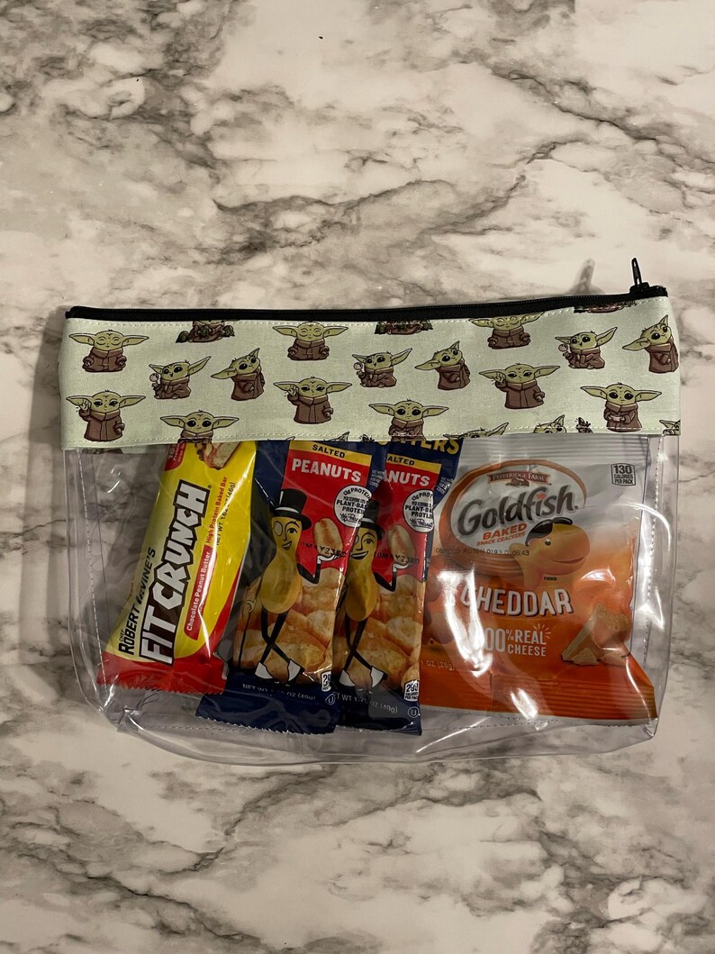 May include: A clear plastic zippered pouch with a green and black fabric lining featuring a repeating pattern of a cartoon character. The pouch contains four snack bags: a red bag of peanut butter crackers, two blue bags of salted peanuts, and a yellow bag of Goldfish crackers.