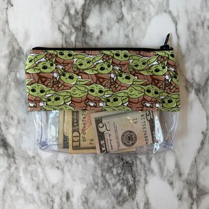 Puede incluir: Una bolsa de pl&aacute;stico transparente con cremallera con un forro de tela verde y marr&oacute;n con un patr&oacute;n repetido de un personaje de dibujos animados. La bolsa est&aacute; abierta y contiene varios billetes de d&oacute;lar estadounidense.