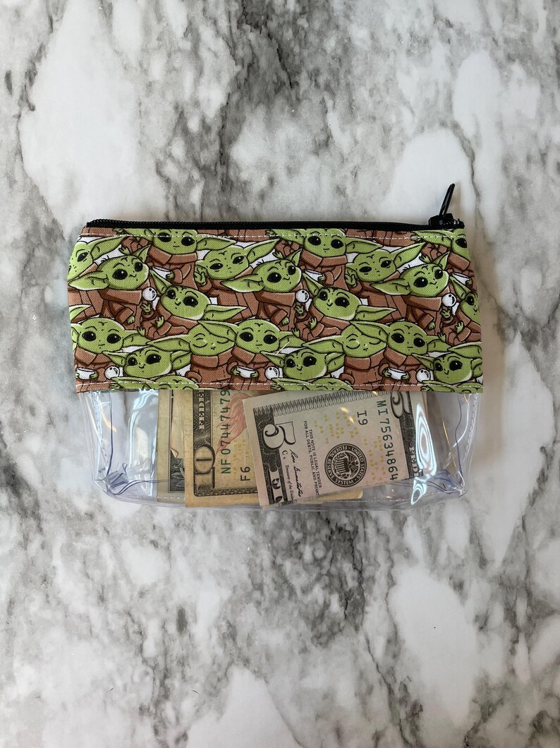 Puede incluir: Una bolsa de pl&aacute;stico transparente con cremallera con un forro de tela verde y marr&oacute;n con un patr&oacute;n repetido de un personaje de dibujos animados. La bolsa est&aacute; abierta y contiene varios billetes de d&oacute;lar estadounidense.