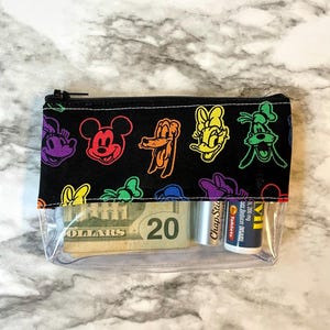 May include: Clear zippered pouch with a black top featuring colorful cartoon character outlines. The pouch contains a twenty-dollar bill, a tube of ChapStick, and a small medicine bottle. The pouch is on a marble-patterned surface.