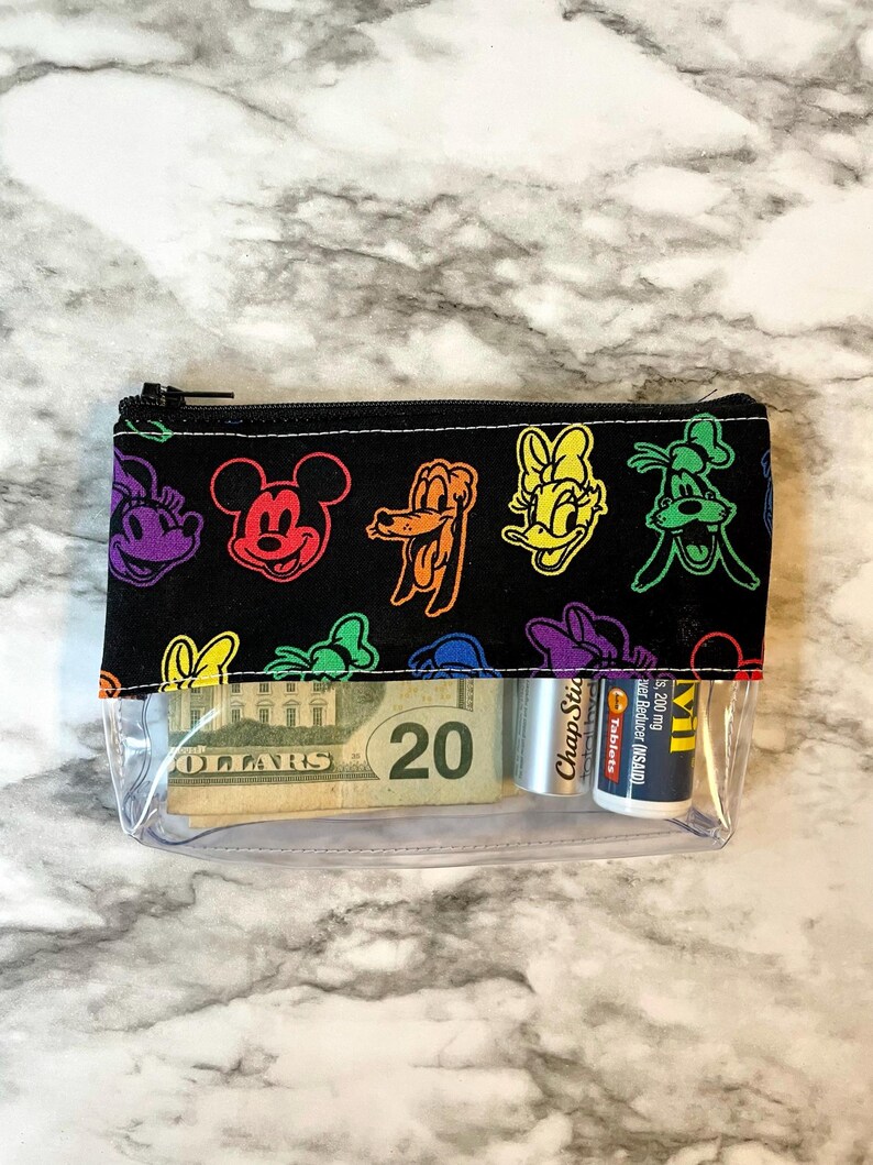 May include: Clear zippered pouch with a black top featuring colorful cartoon character outlines. The pouch contains a twenty-dollar bill, a tube of ChapStick, and a small medicine bottle. The pouch is on a marble-patterned surface.