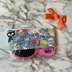 May include: A clear plastic zippered pouch with a blue and white patterned fabric lining. The pouch contains a pink Wet Brush hairbrush. The pouch has a zipper closure and a white trim. The fabric lining has a variety of colorful designs, including flowers, birds, and a black bear. The pouch is sitting on a marble surface.