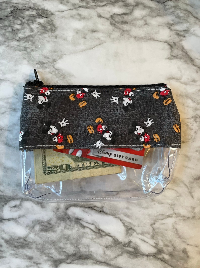 May include: Clear plastic zippered pouch with a black fabric lining featuring a repeating pattern of Mickey Mouse heads. The pouch contains a twenty dollar bill and a Disney gift card.