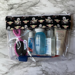 May include: Clear plastic travel bag with a black and green fabric zippered pouch. The pouch has a repeating pattern of a green alien character and a Mandalorian helmet. The bag contains a pink hair brush, a blue toothbrush, a tube of toothpaste, a bottle of hand sanitiser, and a bottle of lotion.