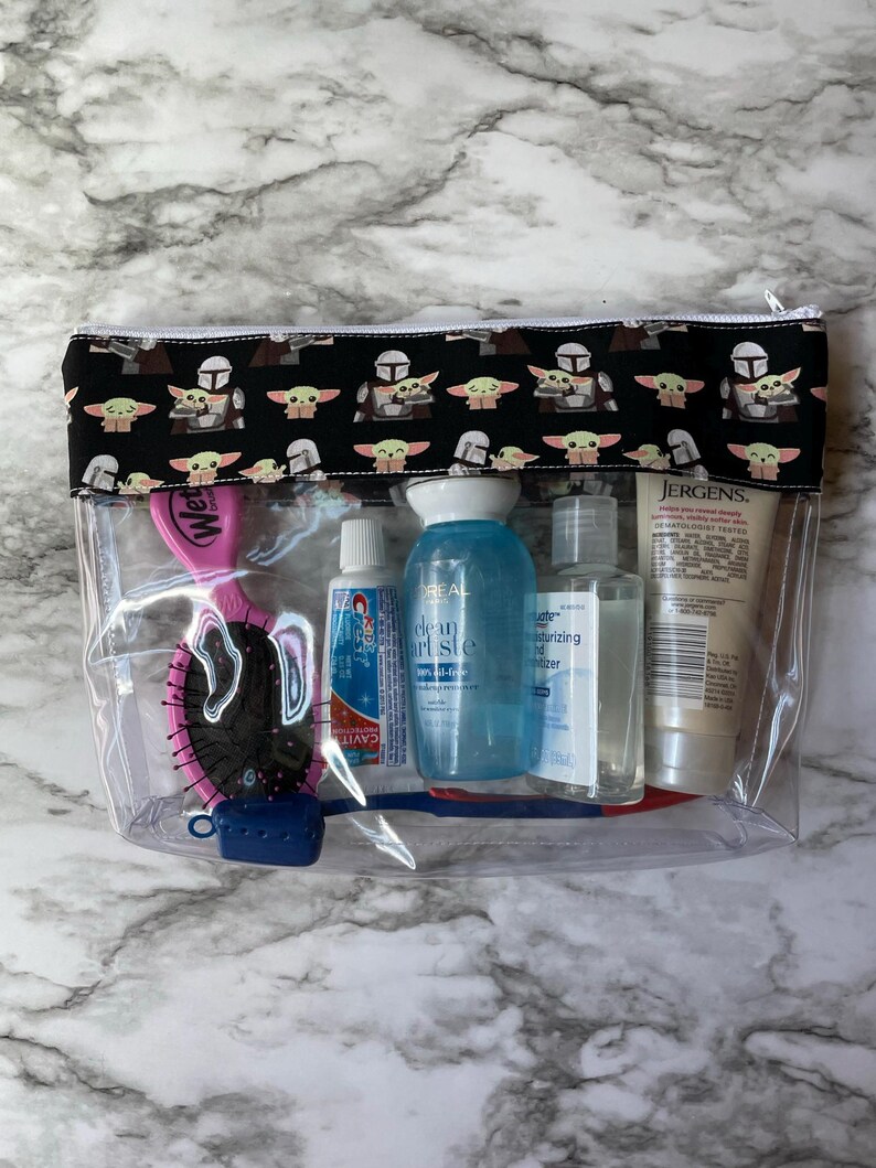 May include: Clear plastic travel bag with a black and green fabric zippered pouch. The pouch has a repeating pattern of a green alien character and a Mandalorian helmet. The bag contains a pink hair brush, a blue toothbrush, a tube of toothpaste, a bottle of hand sanitiser, and a bottle of lotion.