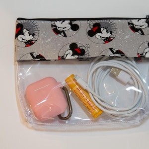 May include: A clear plastic zippered pouch with a grey fabric flap featuring a repeating pattern of Minnie Mouse. The pouch contains a pink case, a yellow lip balm tube, and a white charging cable.