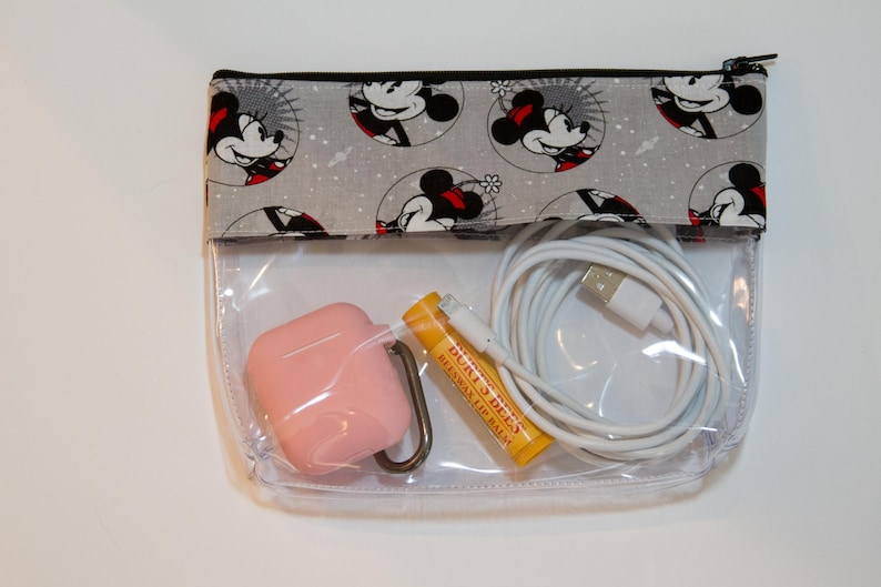 May include: A clear plastic zippered pouch with a grey fabric flap featuring a repeating pattern of Minnie Mouse. The pouch contains a pink case, a yellow lip balm tube, and a white charging cable.
