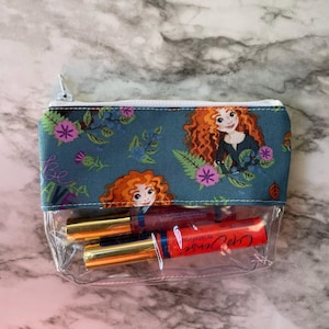 May include: A clear cosmetic bag with a teal fabric top featuring a cartoon illustration of a red-haired character, flowers, and the word "be". The bag contains several lip gloss tubes with gold caps and red labels.