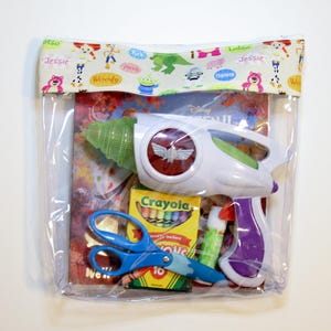 May include: A clear plastic bag with a colorful fabric top featuring characters from the Disney movie Toy Story. The bag contains a toy space ranger blaster, a box of crayons, scissors, and glue.