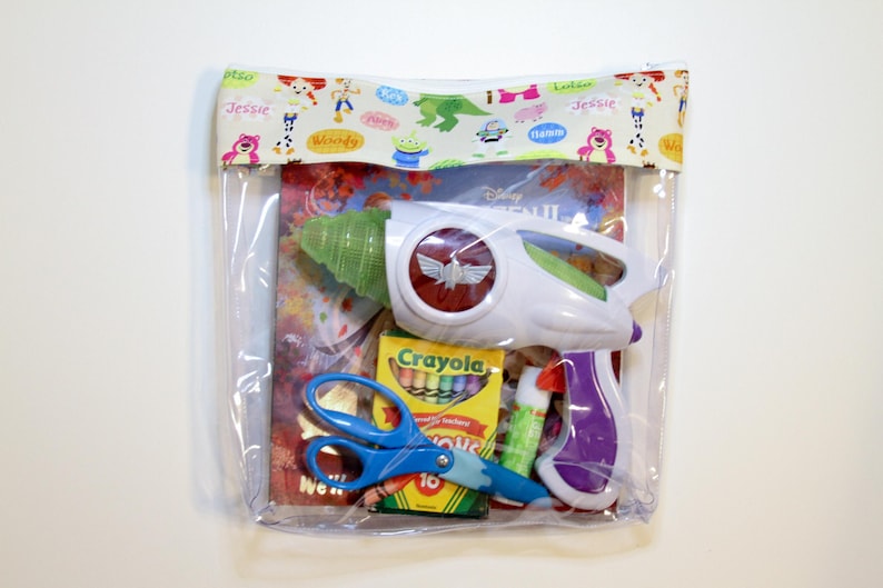 May include: A clear plastic bag with a colorful fabric top featuring characters from the Disney movie Toy Story. The bag contains a toy space ranger blaster, a box of crayons, scissors, and glue.