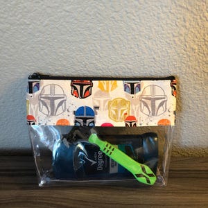 Puede incluir: Una bolsa transparente de pl&aacute;stico con cremallera con un forro de tela blanca que presenta un patr&oacute;n repetido de cascos mandalorianos coloridos. La bolsa contiene un desodorante azul y verde y una maquinilla de afeitar verde.