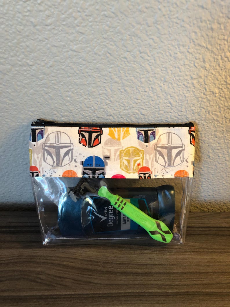 Puede incluir: Una bolsa transparente de pl&aacute;stico con cremallera con un forro de tela blanca que presenta un patr&oacute;n repetido de cascos mandalorianos coloridos. La bolsa contiene un desodorante azul y verde y una maquinilla de afeitar verde.