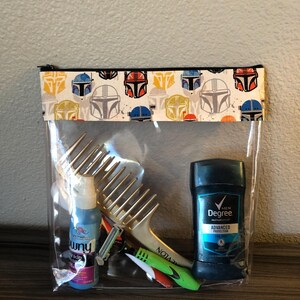 Puede incluir: Una bolsa transparente de pl&aacute;stico con cremallera negra y una solapa de tela estampada. La bolsa contiene un peine dorado, una botella azul de spray, una maquinilla de afeitar verde y roja y un desodorante azul en barra con el texto "MEN Degree ADVANCED PROTECTION" en la etiqueta.