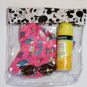 K&ouml;nnte beinhalten: Eine transparente Reisetasche mit Rei&szlig;verschluss und einem Mickey Mouse-Druck am oberen Rand. Im Inneren befinden sich ein rosa Sonnenhut, eine Sonnenbrille und eine gelbe Sonnencreme-Flasche. Der Hut hat ein farbenfrohes Muster.