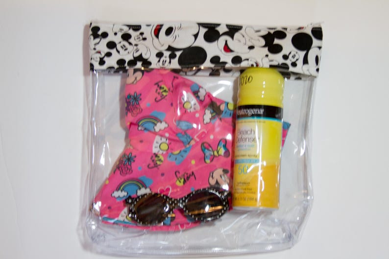 K&ouml;nnte beinhalten: Eine transparente Reisetasche mit Rei&szlig;verschluss und einem Mickey Mouse-Druck am oberen Rand. Im Inneren befinden sich ein rosa Sonnenhut, eine Sonnenbrille und eine gelbe Sonnencreme-Flasche. Der Hut hat ein farbenfrohes Muster.