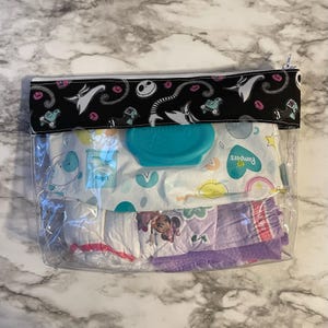 May include: A clear plastic zippered pouch with a black and white fabric lining featuring a cartoon character. The pouch contains a package of baby wipes, a container of baby wipes, and a package of training pants with a cartoon character on them.