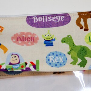 May include: A white zippered pouch with a clear plastic front. The pouch is decorated with a colorful pattern of Toy Story characters, including Woody, Buzz Lightyear, Rex, and Alien. The text "Bullseye", "Rex", "Alien", and "Lots-o-Huggin' Bear" is printed on the pouch.