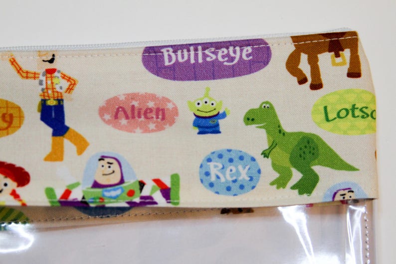 May include: A white zippered pouch with a clear plastic front. The pouch is decorated with a colorful pattern of Toy Story characters, including Woody, Buzz Lightyear, Rex, and Alien. The text "Bullseye", "Rex", "Alien", and "Lots-o-Huggin' Bear" is printed on the pouch.
