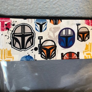 Puede incluir: Una bolsa de cremallera de tela blanca con un frente de pl&aacute;stico transparente. La tela tiene un patr&oacute;n repetido de cascos Mandalorianos coloridos.