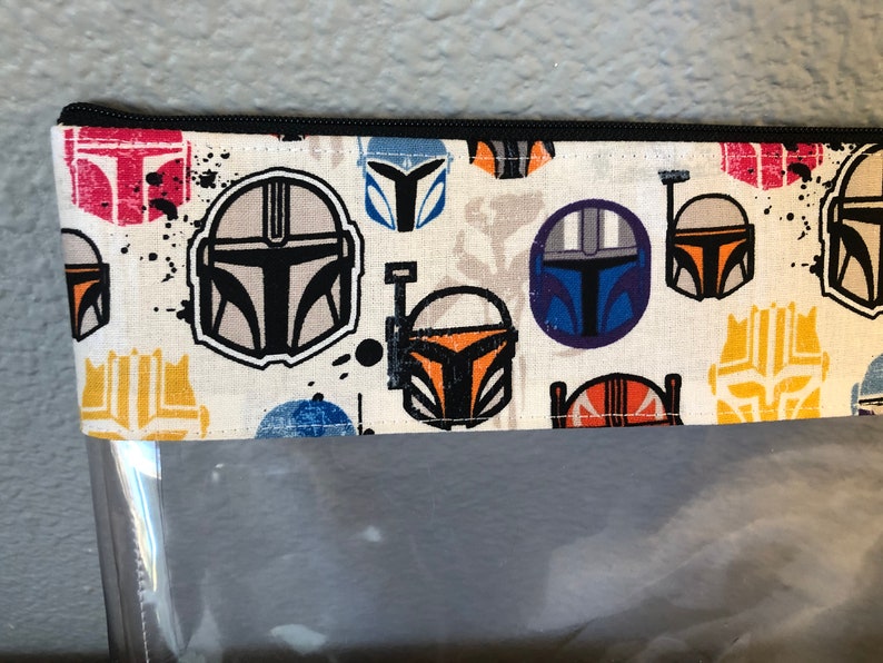 Puede incluir: Una bolsa de cremallera de tela blanca con un frente de pl&aacute;stico transparente. La tela tiene un patr&oacute;n repetido de cascos Mandalorianos coloridos.