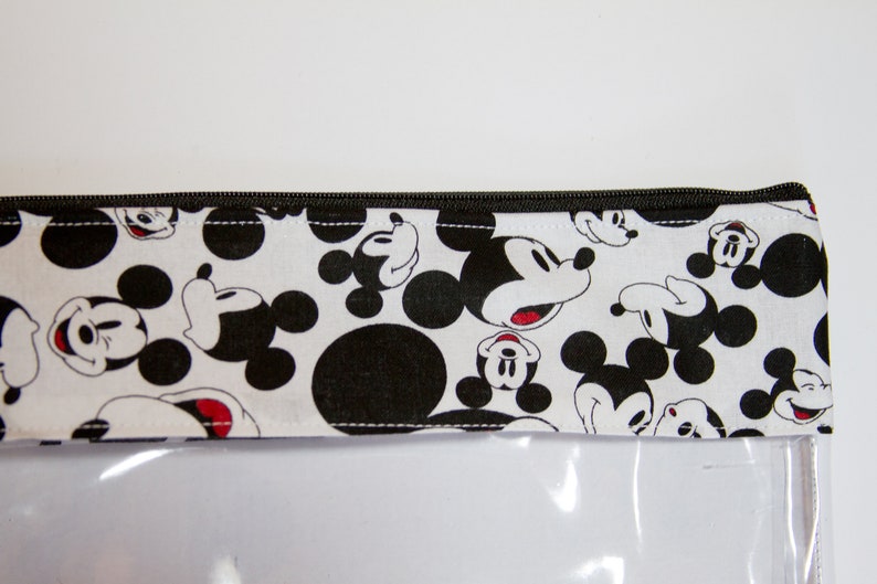 K&ouml;nnte beinhalten: Eine transparente, rechteckige Tasche mit einem schwarzen Rei&szlig;verschluss und einer wei&szlig;en Stoffoberseite. Der Stoff ist mit schwarz-wei&szlig;en Mickey Mouse-Gesichtern und Tupfen gemustert. Die Tasche ist wahrscheinlich zur Aufbewahrung.