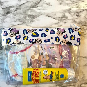 Pode incluir: Uma bolsa transparente com fecho de correr de pl&aacute;stico com fundo branco e estampado de Minnie Mouse. A bolsa cont&eacute;m uma fralda, um tubo de creme para assaduras e uma garrafa de gel de banho para beb&eacute;s. O tubo de creme para assaduras diz "ORIGINAL BOUDREAUX'S BUTT PASTE DIAPER RASH OINTMENT IN PROTECTANT (ZINC OXIDE)". A garrafa de gel de banho para beb&eacute;s diz "Trata e previne assaduras Sem parabenos e sem conservantes".