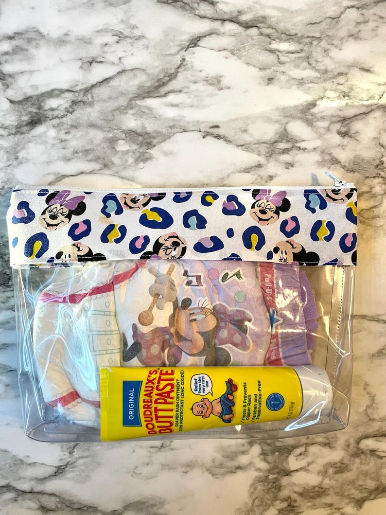Pode incluir: Uma bolsa transparente com fecho de correr de pl&aacute;stico com fundo branco e estampado de Minnie Mouse. A bolsa cont&eacute;m uma fralda, um tubo de creme para assaduras e uma garrafa de gel de banho para beb&eacute;s. O tubo de creme para assaduras diz "ORIGINAL BOUDREAUX'S BUTT PASTE DIAPER RASH OINTMENT IN PROTECTANT (ZINC OXIDE)". A garrafa de gel de banho para beb&eacute;s diz "Trata e previne assaduras Sem parabenos e sem conservantes".