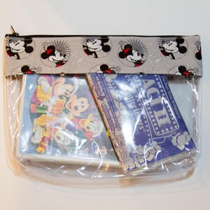 May include: A clear plastic zipped pouch with a grey and white fabric flap featuring a repeating pattern of Mickey Mouse heads. The pouch contains two items: a blue and white card with the text "ACT II Butter Lovers" and a clear plastic case with a cartoon image of Mickey Mouse and friends.