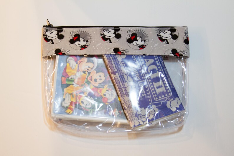 May include: A clear plastic zipped pouch with a grey and white fabric flap featuring a repeating pattern of Mickey Mouse heads. The pouch contains two items: a blue and white card with the text "ACT II Butter Lovers" and a clear plastic case with a cartoon image of Mickey Mouse and friends.