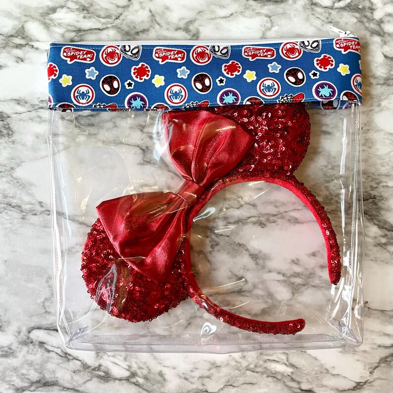May include: A clear plastic zippered pouch with a red and blue patterned fabric flap. The pouch contains a pair of red sequined Minnie Mouse ears with a red satin bow.