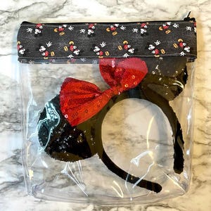 May include: A clear plastic zippered pouch with a black fabric lining featuring a repeating pattern of Mickey Mouse and Minnie Mouse. Inside the pouch is a black headband with red sequin ears and a red bow.