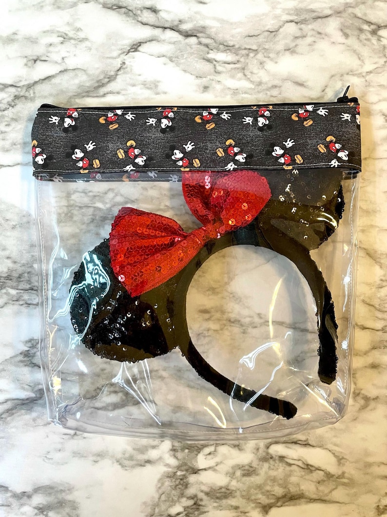 May include: A clear plastic zippered pouch with a black fabric lining featuring a repeating pattern of Mickey Mouse and Minnie Mouse. Inside the pouch is a black headband with red sequin ears and a red bow.