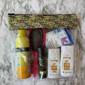 Puede incluir: Una bolsa transparente con cremallera de pl&aacute;stico con un estampado verde y marr&oacute;n de un personaje de dibujos animados. La bolsa contiene una botella de laca para el cabello amarilla, un cepillo para el cabello rosa, una botella blanca de acondicionador para el cabello, una botella blanca de champ&uacute; y una lata de laca para el cabello negra. El texto en las botellas dice "Pantene Moisture" y "TRESemme Botanique Nourish & Replenish".