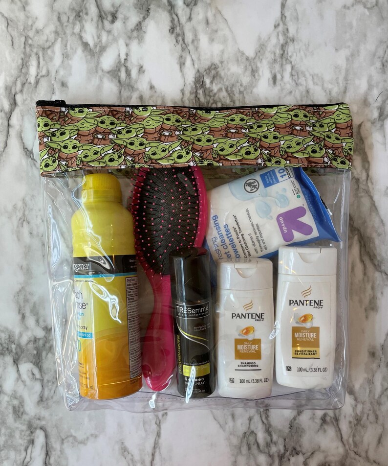 Puede incluir: Una bolsa transparente con cremallera de pl&aacute;stico con un estampado verde y marr&oacute;n de un personaje de dibujos animados. La bolsa contiene una botella de laca para el cabello amarilla, un cepillo para el cabello rosa, una botella blanca de acondicionador para el cabello, una botella blanca de champ&uacute; y una lata de laca para el cabello negra. El texto en las botellas dice "Pantene Moisture" y "TRESemme Botanique Nourish & Replenish".