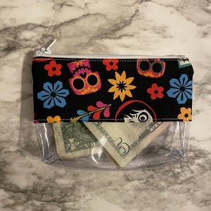 May include: A clear plastic zippered pouch with a black fabric lining. The fabric has a colorful pattern of sugar skulls, flowers, and arrows. The pouch is open and contains two US dollar bills.