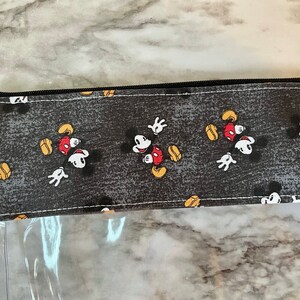 May include: A clear plastic zippered pouch with a black zipper and a gray fabric flap featuring a repeating pattern of Mickey Mouse in various poses.