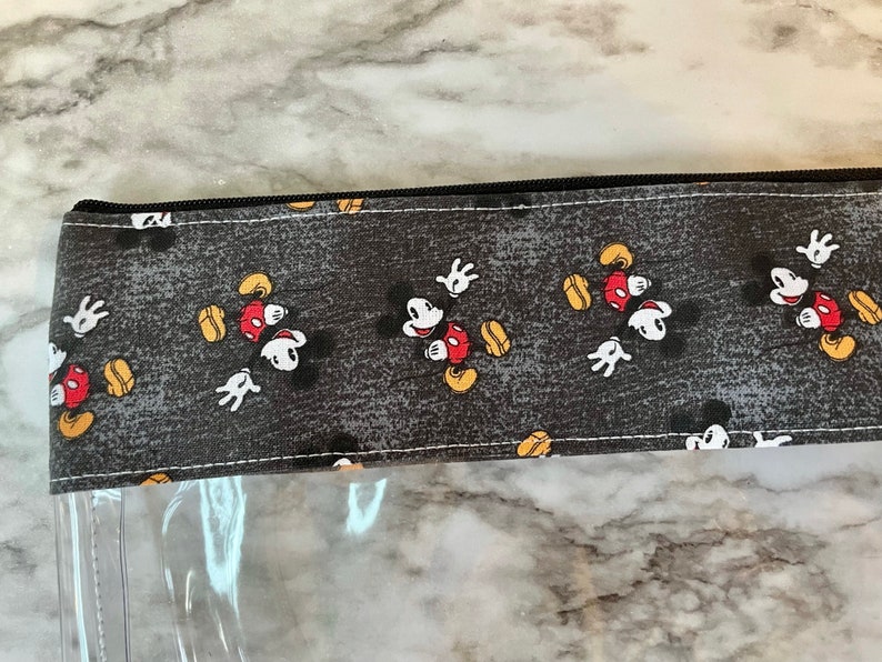 May include: A clear plastic zippered pouch with a black zipper and a gray fabric flap featuring a repeating pattern of Mickey Mouse in various poses.
