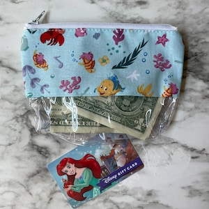 May include: A clear plastic zippered pouch with a blue and white fabric lining. The fabric has a pattern of sea creatures, including a crab, a fish, and a starfish. The pouch is open and contains a few dollar bills and a Disney gift card featuring Ariel from The Little Mermaid.